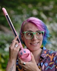 Valerie, who has brightly colored pink and blue hair and mint green glasses, is smiling while looking off to the side. They are looking off to the side and holding a Kirby otomatone which is a silly musical instrument.